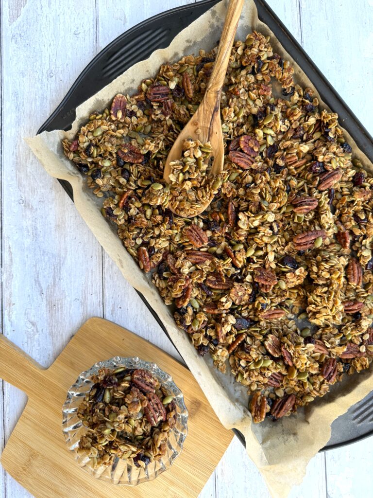 Wild blueberry granola presented on a sheet pan beside a wood cutting board with a serving of the granola in a clear bowl.