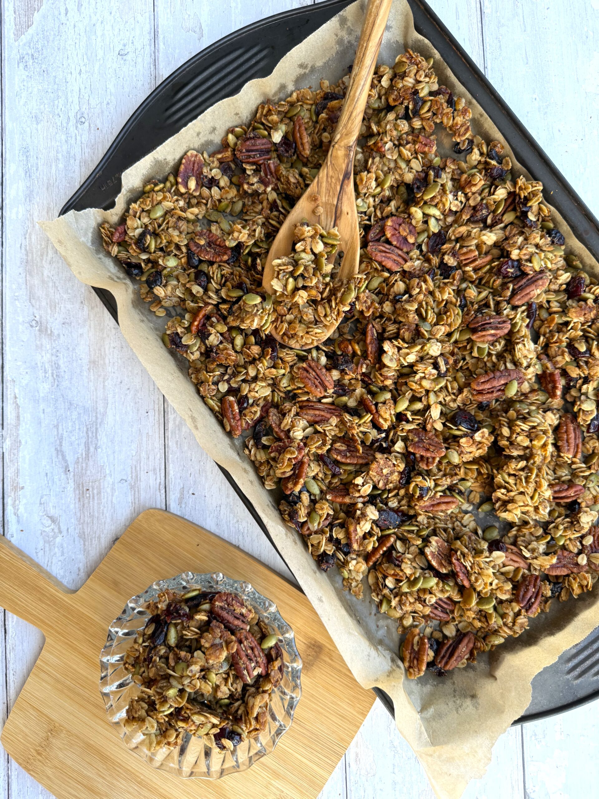 Wild blueberry granola presented on a sheet pan beside a wood cutting board with a serving of the granola in a clear bowl.