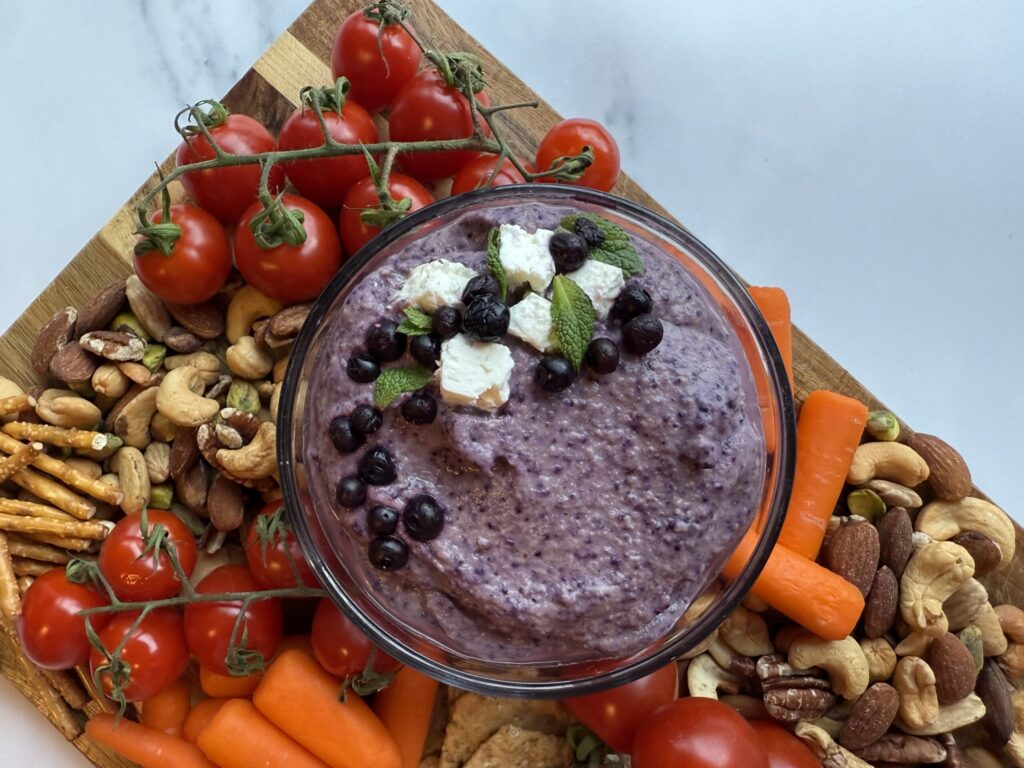 Wild blueberry hummus in a clear bowl along side carrots, nuts, pretzels sitting on a wood cutting board.