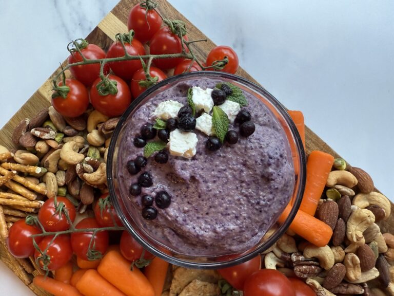 Wild blueberry hummus in a clear bowl along side carrots, nuts, pretzels sitting on a wood cutting board.